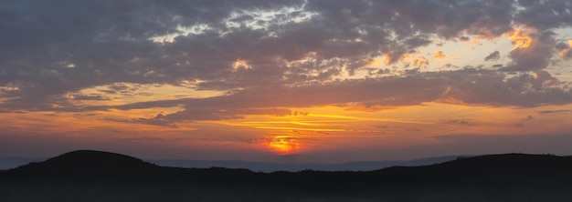 Panorama eines dramatischen farbenfrohen Sonnenuntergangs mit dunklen und hellen Wolken vor dem Hintergrund der Berge. Berge im Nebel auf dem Hintergrund des Sonnenuntergangs. Die Sonnenstrahlen bahnen sich ihren Weg durch die cl