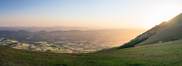 Panorama-Luftbild bei Sonnenuntergang in der Region Umbrien, Italien. Sommergrünes Tal einzigartige Hügel- und Berglandschaft. Bebaute Felder in der Ebene darunter.