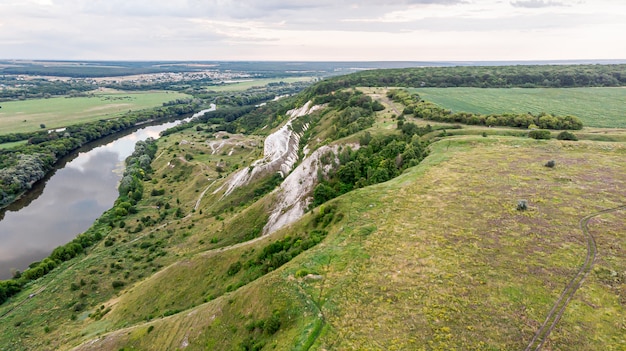 Panorama-Luftbild über der Spitze einer Sommerlandschaft eines grünen landwirtschaftlichen Feldes, der Hügel oder der Berge, eines großen Flusses und eines Waldgürtels bei Sonnenuntergang
