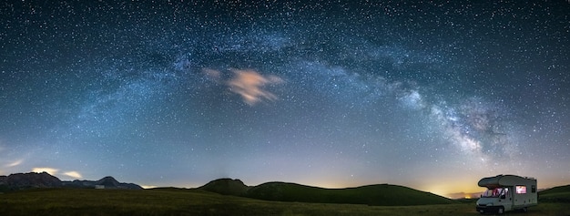 Foto panorama nachthimmel über campo imperatore hochland, abruzzen, italien. der bogen der milchstraße und die sterne über einem beleuchteten wohnmobil. campingfreiheit in einzigartiger hügellandschaft.