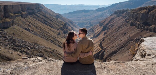 Panorama de la pareja romántica en el amor abraza y mira a los ojos con gafas de sol y sentado en el borde del acantilado empinado contra el paisaje de montaña