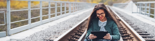 Panorama-perspektivisches Porträt des lächelnden Teenager-Mädchens, das auf Eisenbahn sitzt und digitales intelligentes Tablett verwendet. Brille und grüne Jacke tragen.