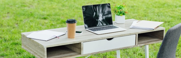 Panoramaaufnahme eines weißen Tisches mit Laptop, Smartphone, Coffee-to-go-Notizbüchern und Pflanze im Park