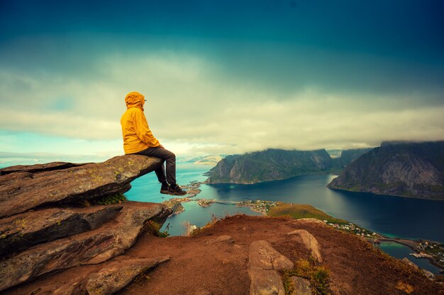 Panoramablick auf den Fjord und das Fischerdorf. Manntourist, der auf einer Felsenklippe sitzt. Schöne Berglandschaft. Natur Norwegen, Lofoten-Inseln.