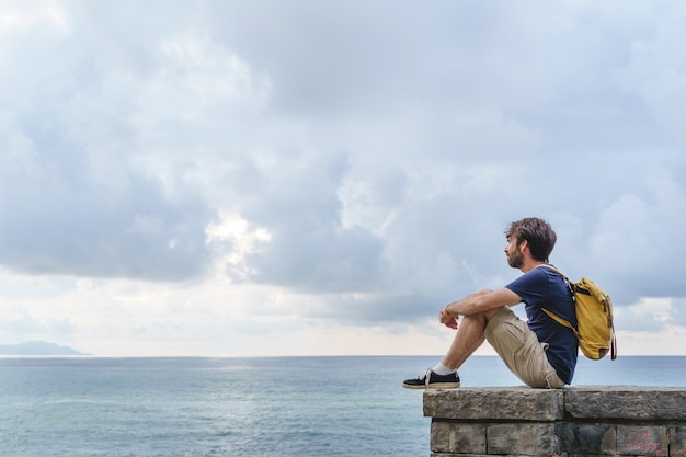 Panoramablick auf den jungen Reisenden Mann sitzt in einer Klippe. Horizontale Ansicht von Rucksacktouristen, die im Freien mit dem blauen Ozean im Hintergrund reisen.