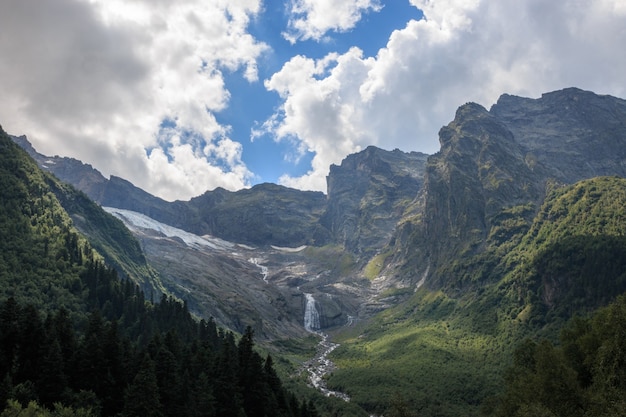 Panoramablick auf die Berge und den weit entfernten Wasserfall im Nationalpark von Dombay, Kaukasus, Russland. Sommerlandschaft, Sonnenscheinwetter und sonniger Tag