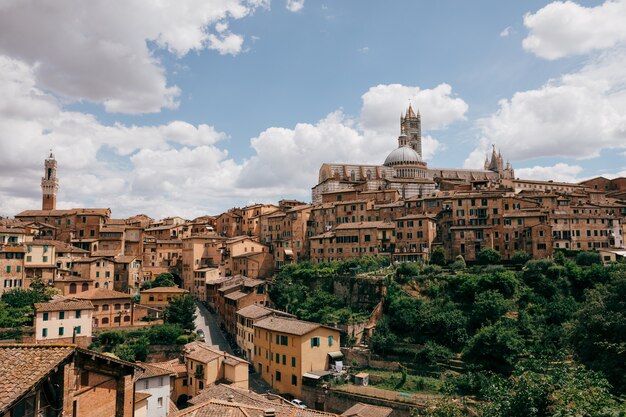 Panoramablick auf die Stadt Siena mit historischen Gebäuden und dem weit entfernten Dom von Siena (Duomo di Siena). Sonniger Sommertag und dramatischer blauer Himmel