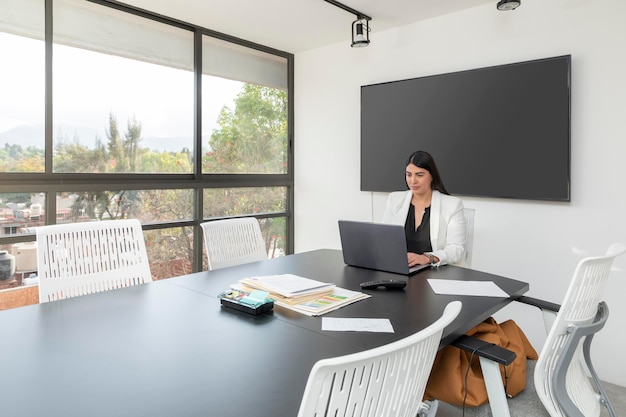 Panoramablick auf eine leitende Frau, die in ihrem Büro mit ihrem Laptop und einem Bildschirm für Präsentationen an der Wand und Fenstern im Hintergrund mit Blick nach draußen arbeitet