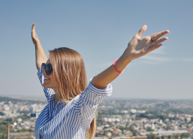 Panoramablick auf eine Stadt und eine junge Frau mit Sonnenbrille im Vordergrund, die ihre Arme hochhält