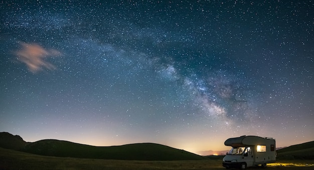 Panorámica del cielo nocturno sobre las tierras altas de Campo Imperatore, Abruzzo, Italia. El arco de la galaxia de la Vía Láctea y las estrellas sobre una caravana iluminada. Libertad de acampada en un paisaje de colinas único.