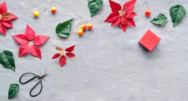 Panorámica de Navidad, pancarta sobre tela de lino natural. Poinsettia roja, decoración navideña, manzanas silvestres naranjas y caja de regalo roja. Tijeras, poinsettia rojo y verde oscuro hojas sobre mantel beige.
