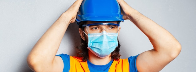 Panorámica retrato de banner de joven confiado con máscara médica en la cara, ingeniero trabajador de la construcción con equipo de seguridad sobre fondo texturizado de gris. Prevención contra el coronavirus.