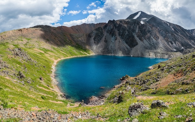 Panoramische Alpenlandschaft mit Bergsee im grünen Tal und Gletscher unter bewölktem Himmel. Atmosphärische Hochlandlandschaft mit wunderschönem Gletschersee zwischen sonnenbeschienenen Hügeln und Felsen gegen Bergkette