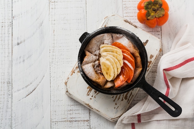 Panqueca de bebê holandês com maçã, caqui, banana, canela em uma pequena panela de ferro na superfície da mesa de madeira. Vista do topo.