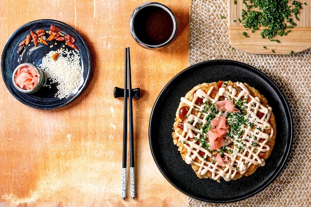 Panqueca de repolho de okonomiyaki de fast food japonesa caseira decorada com cebolinha, gengibre em conserva, molho de maionese em prato de cerâmica preta com pauzinhos e ingredientes acima. Mesa de textura. Postura plana