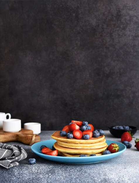 Panquecas caseiras em um prato azul com frutas e xarope em um fundo de concreto.