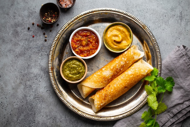 Panquecas de arroz indiano tradicional Dosa com diferentes molhos chutney e temperos na placa de metal rústica na mesa de fundo de pedra. Refeição rápida ou lanche vegetariano do sul da Índia, vista de cima