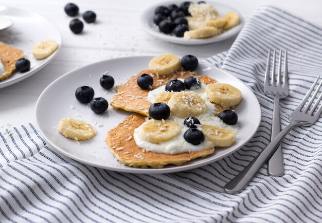 Panquecas de aveia caseiro com iogurte, mirtilo fresco e banana na mesa de madeira branca.