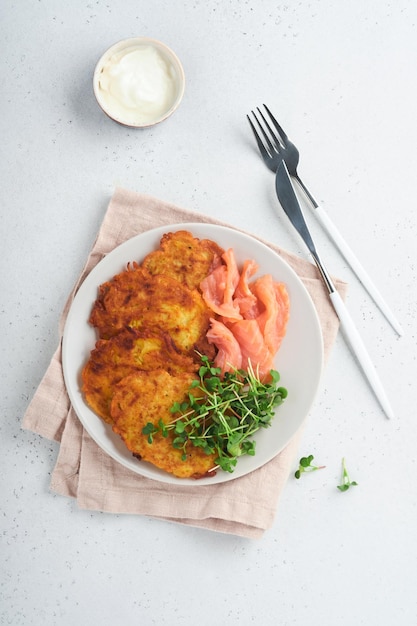 Panquecas de batata Panquecas de batata caseira frita ou latkes com creme de cebola verde microgreens salmão vermelho e molho em prato rústico no fundo da mesa branca Estilo rústico Comida saudável Vista superior