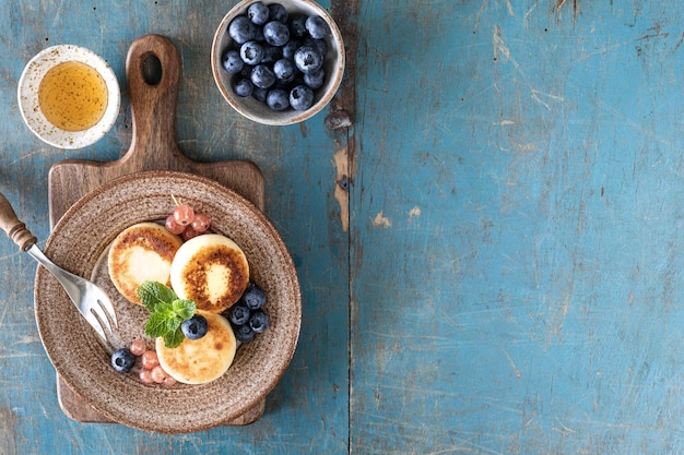 Panquecas de queijo cottage, cheesecakes, bolinhos de ricota com mirtilos frescos, groselhas e pêssegos em um prato Café da manhã saudável e delicioso para o feriado Fundo de madeira azul