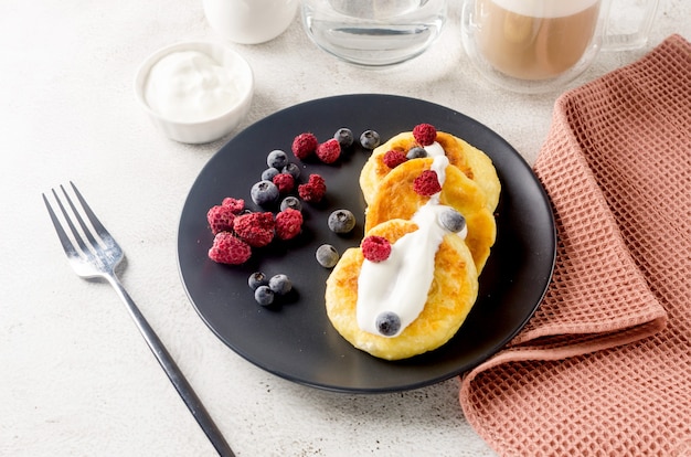 Panquecas de queijo cottage em uma placa preta com baga, creme, geléia e xícara de café com leite. Café da manhã saudável. Comida caseira