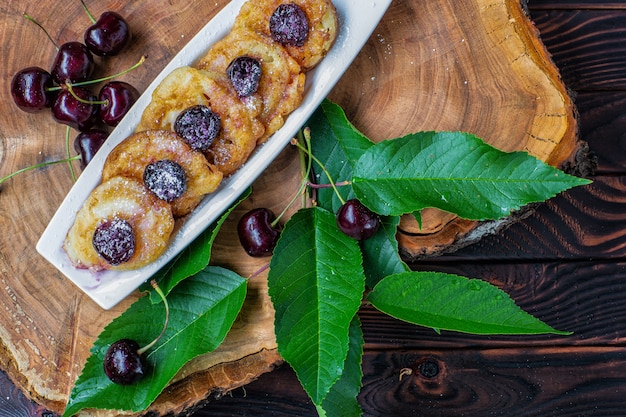 Panquecas em molho de cereja em um prato e bagas de cereja com folhas verdes em uma placa de madeira