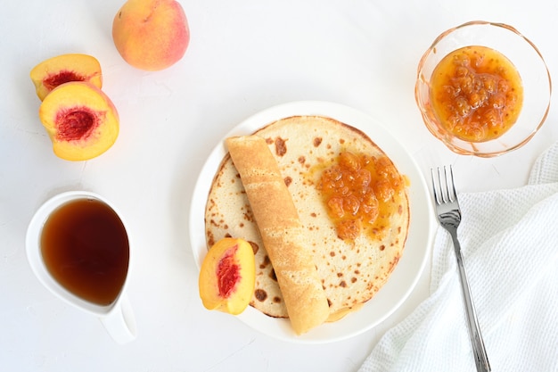 Panquecas grandes com geléia, chá e pêssegos em fundo branco.