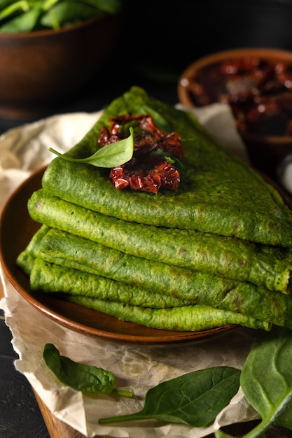 Panquecas vegetarianas verdes decoradas com tomates secos. Panquecas de espinafre em um prato marrom no fundo escuro de madeira.