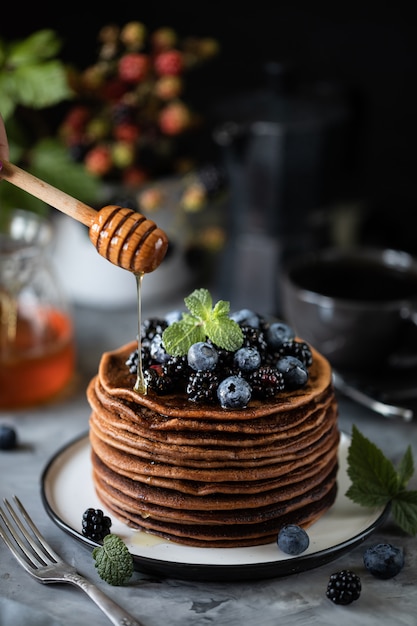 Panqueque de chocolate con bayas y frutas con miel, con azúcar en polvo volador y un ramo de flores silvestres sobre la mesa. Oscuro