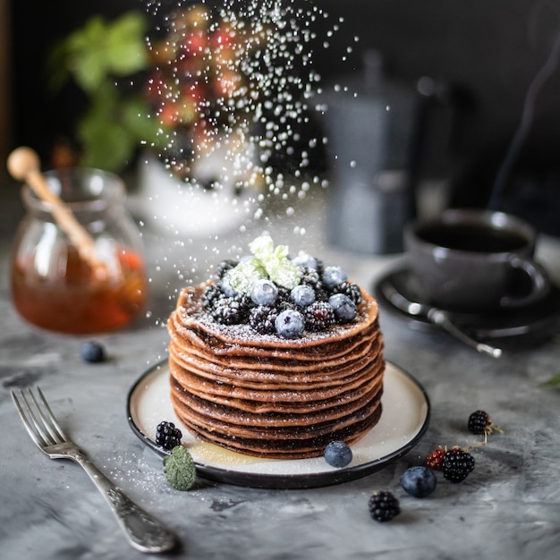 Panqueque de chocolate con bayas y frutas con miel, con azúcar en polvo volador y un ramo de flores silvestres sobre la mesa. Oscuro