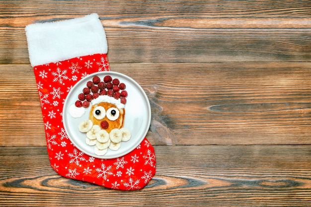 Panqueque en forma de cara de Papá Noel de Navidad con baya de frambuesa fresca dulce, plátano en placa, calcetín de Navidad sobre fondo de madera para el desayuno de los niños de los niños. postre de comida navideña, decoraciones de año nuevo