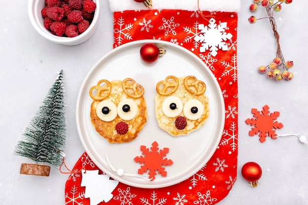 Foto panqueque con forma de cara de venado navideño con dulce de frambuesa fresca y queso en un plato para niños, bebés, niños, desayuno, cena, comida de navidad con adornos de año nuevo sobre fondo gris de cerca