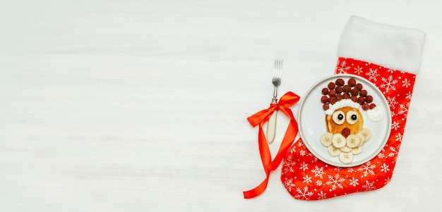 Panqueque de Navidad con forma de Papá Noel con bayas de frambuesa fresca dulce y plátano en placa sobre fondo blanco de madera para el desayuno de los niños. Decoraciones de año nuevo de comida de Navidad con banner de espacio de copia.