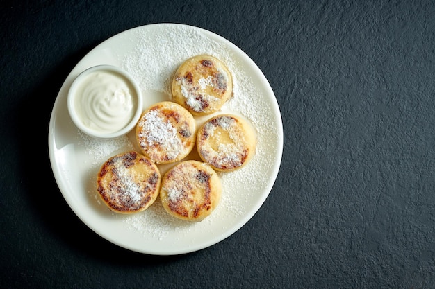 Panqueque de tarta de queso con crema agria en un plato blanco sobre un fondo negro. Sírniki