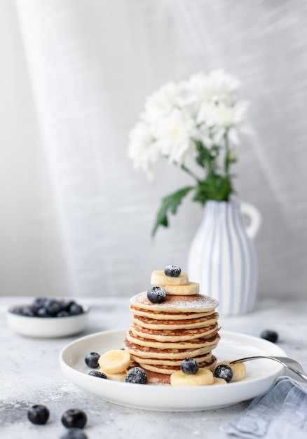 Panqueques con arándanos y plátano en una mesa con flores en un florero. concepto de desayuno saludable imagen vertical