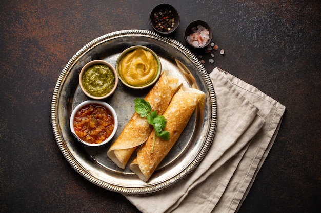 Panqueques de arroz indio tradicional Dosa con diferentes salsas chutney, condimentos en placa de metal rústico en la mesa de fondo de piedra marrón oscuro. Comida rápida o bocadillo vegetariano del sur de la India, vista superior