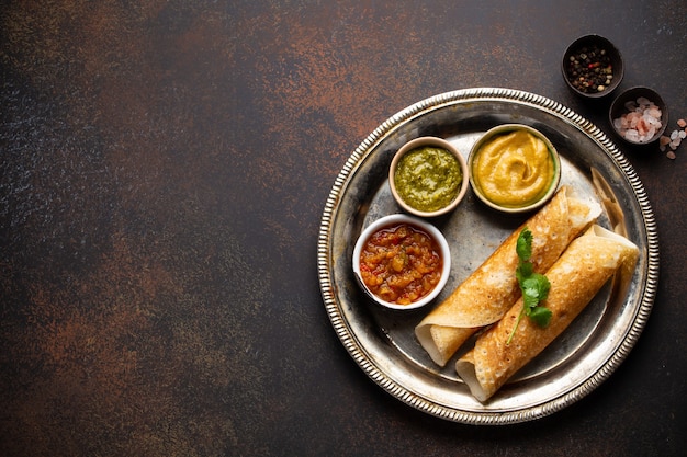 Panqueques de arroz indio tradicional Dosa con diferentes salsas chutney, sobre placa de metal rústico sobre tabla de fondo de piedra marrón oscuro. Comida rápida o refrigerio vegetariano del sur de la India, vista superior, espacio para texto