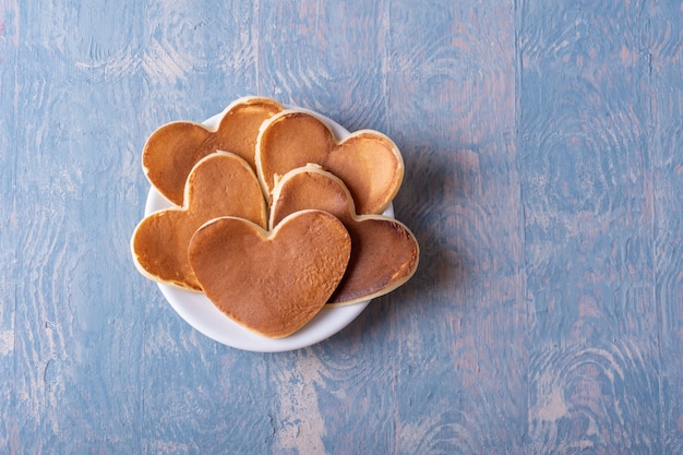 Panqueques caseros en forma de corazón en un plato blanco sobre una mesa de madera azul, copie el espacio, vista superior.