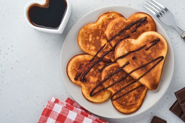 Panqueques en forma de corazones de desayuno con salsa de chocolate en placa de cerámica gris taza de café sobre fondo de hormigón gris. Ajuste de la tabla para su desayuno favorito del Día de San Valentín Vista superior del espacio de copia