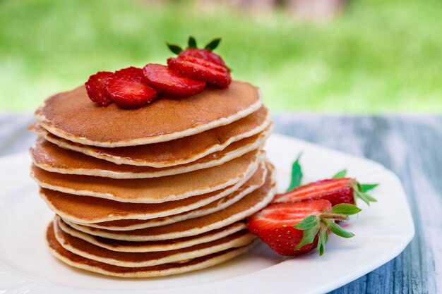 Panqueques con fresas frescas y menta en plato blanco en jardín de madera rosa o en la naturaleza. Pila de panqueques en la mesa.