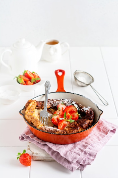 Panqueques holandeses para bebés con fresas frescas y espolvoreados con azúcar glas en polvo en una sartén roja sobre la superficie blanca de la cocina. Vista superior.