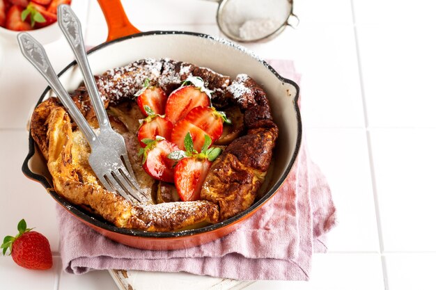 Panqueques holandeses para bebés con fresas frescas y espolvoreados con azúcar glas en polvo en una sartén roja sobre la superficie blanca de la cocina. Vista superior.