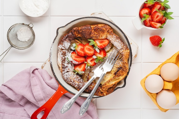 Panqueques holandeses para bebés con fresas frescas y espolvoreados con azúcar en polvo en una sartén roja sobre la pared blanca de la cocina. Vista superior.