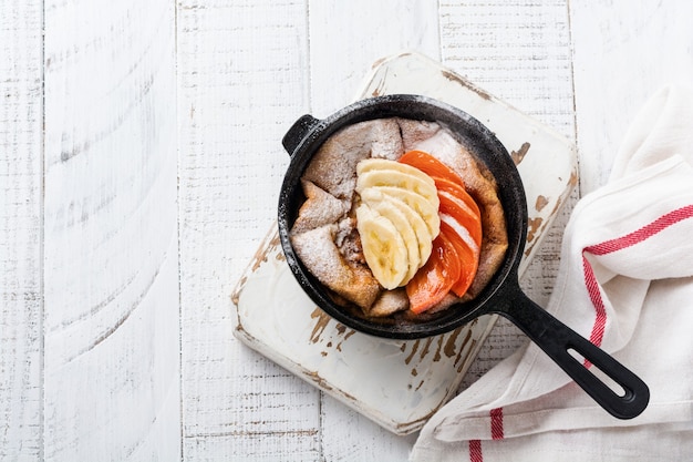 Panqueques holandeses para bebés con manzana, caqui, plátano, canela en una sartén de hierro pequeña sobre la superficie de la mesa de madera. Vista superior.