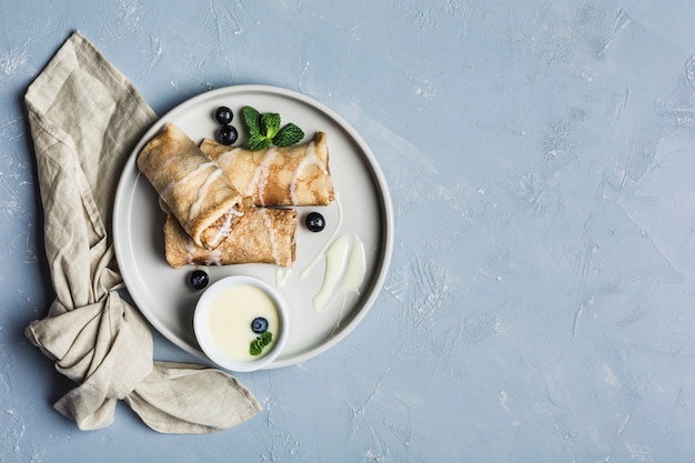 Unos panqueques con relleno en una placa gris con arándanos y menta, con leche dulce condensada en una salsera sobre un fondo azul claro.