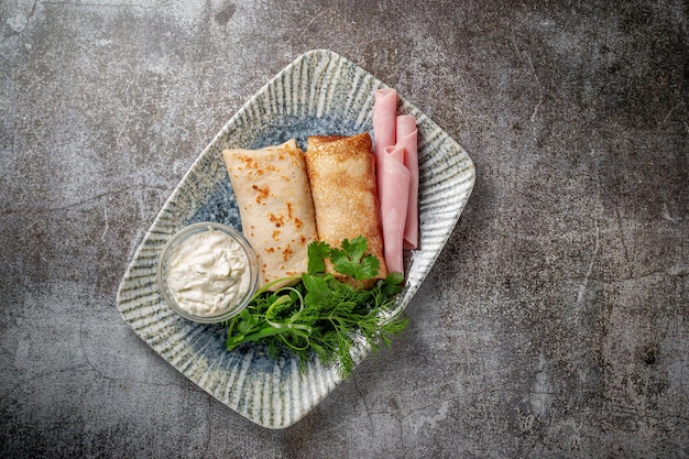 Panqueques rellenos de jamón en un plato con perejil y hojas de eneldo y crema agria sobre una mesa de piedra gris, flatlay