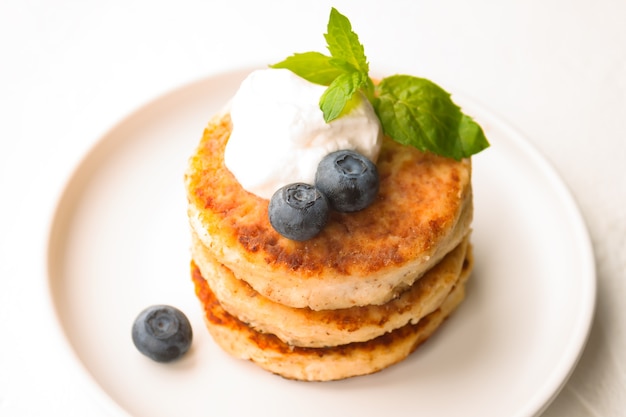 Panqueques de requesón o buñuelos de cuajada se apilan con crema agria, hojas de menta y arándanos de almendra y harina de coco sin gluten en la vista cercana de la placa blanca. Concepto de desayuno. Enfoque suave selectivo.