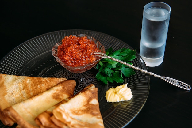 Panqueques rusos dorados con caviar rojo y mantequilla en un plato transparente cerca del vaso de vodka de hielo