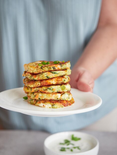 Panqueques de verduras o buñuelos apilados en un plato en la mano de una mujer fuera de foco un tazón con salsa de yogur sobre la mesa