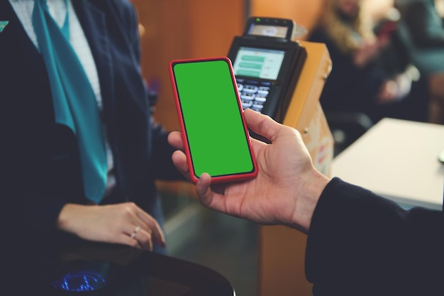 Pantalla en blanco de la llave de croma verde del teléfono inteligente con espacio publicitario para aplicaciones móviles en una mano cerca de la terminal bancaria mientras pasa por el tablero de facturación del vuelo